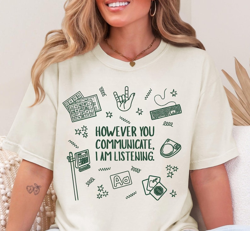 May include: Cream-colored t-shirt with the text "HOWEVER YOU COMMUNICATE, I AM LISTENING." and various green illustrations, including a keyboard, hand sign, and cards. The shirt has a relaxed fit.