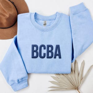 Könnte beinhalten: Ein hellblaues Sweatshirt mit Rundhalsausschnitt und den Buchstaben BCBA, die in dunkelblauem Garn mittig auf der Brust aufgestickt sind. Das Kleidungsstück hat lange Ärmel, einen gerippten Halsausschnitt und eine Standardpassform mit Etikett.