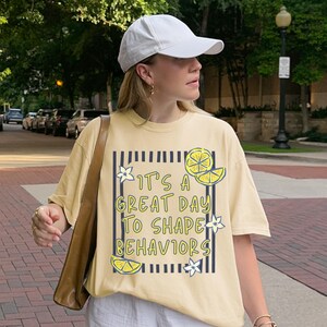 May include: A light yellow t-shirt with the text "It's a great day to shape behaviors" in a black and yellow design. The shirt features lemon and flower graphics. The person in the image is wearing a white baseball cap.