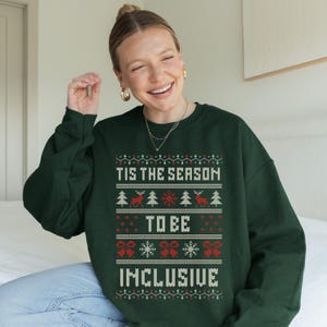 May include: A dark green crewneck sweatshirt featuring a white and red holiday-themed knit pattern. The graphic includes the text 'TIS THE SEASON TO BE INCLUSIVE' surrounded by motifs of reindeer, pine trees, snowflakes, and bows in a festive style.