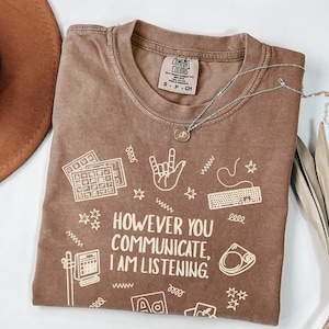 May include: Brown Comfort Colors t-shirt with the text "HOWEVER YOU COMMUNICATE, I AM LISTENING." and various white graphics. A silver necklace with a pendant is on the shirt. A brown hat is visible on the left.