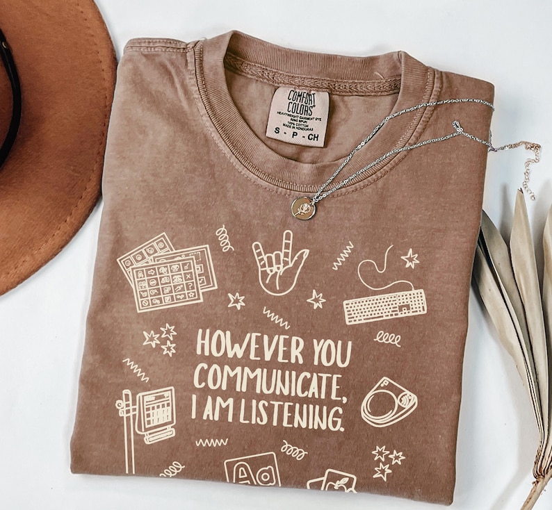 May include: Brown Comfort Colors t-shirt with the text "HOWEVER YOU COMMUNICATE, I AM LISTENING." and various white graphics. A silver necklace with a pendant is on the shirt. A brown hat is visible on the left.