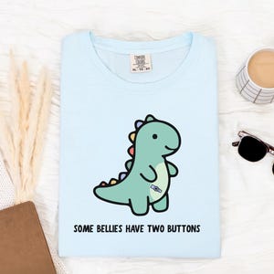 May include: A pale blue t-shirt with a cartoon dinosaur illustration. The dinosaur is green with a button detail and colourful spines. The text "SOME BELLIES HAVE TWO BUTTONS" is printed below. The shirt is a Comfort Colors XL-TG-EG.