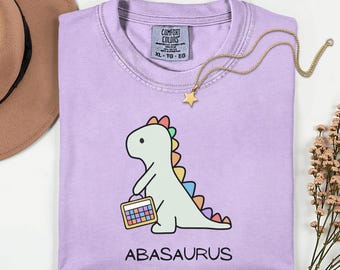 May include: A light purple t-shirt featuring a cartoon dinosaur with a rainbow spine and carrying a colorful calculator. The word "ABASAURUS" is printed below the dinosaur. A gold necklace with a star pendant is draped on the shirt.