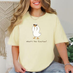 K&ouml;nnte beinhalten: Hellgelbes T-Shirt mit einer Cartoon-Ente, die eine Sonnenbrille tr&auml;gt. Die Ente ist wei&szlig; mit orangefarbenen F&uuml;&szlig;en und einem schwarzen Umriss. Der Text "What's the function?" ist darunter gedruckt.