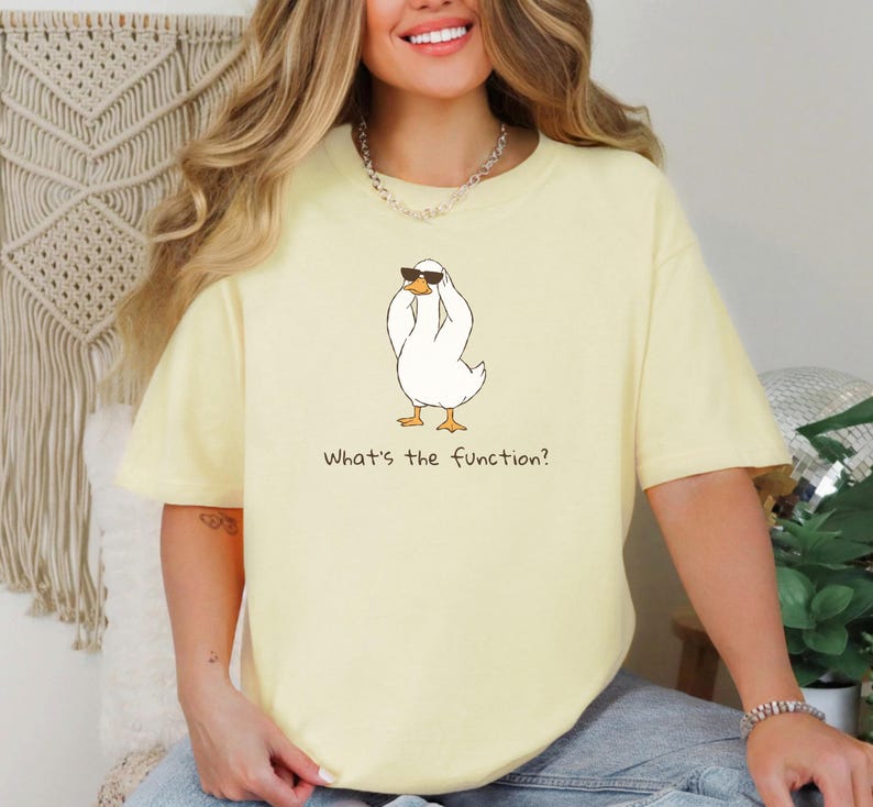 K&ouml;nnte beinhalten: Hellgelbes T-Shirt mit einer Cartoon-Ente, die eine Sonnenbrille tr&auml;gt. Die Ente ist wei&szlig; mit orangefarbenen F&uuml;&szlig;en und einem schwarzen Umriss. Der Text "What's the function?" ist darunter gedruckt.