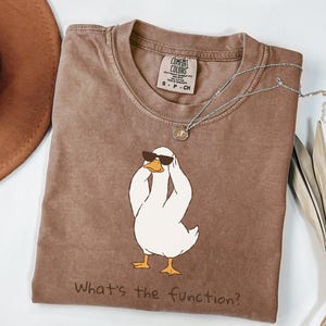 May include: A brown Comfort Colors t-shirt with a white duck wearing sunglasses and holding its head. The text "What's the function?" is printed below the duck. A silver necklace with a pendant is also visible.