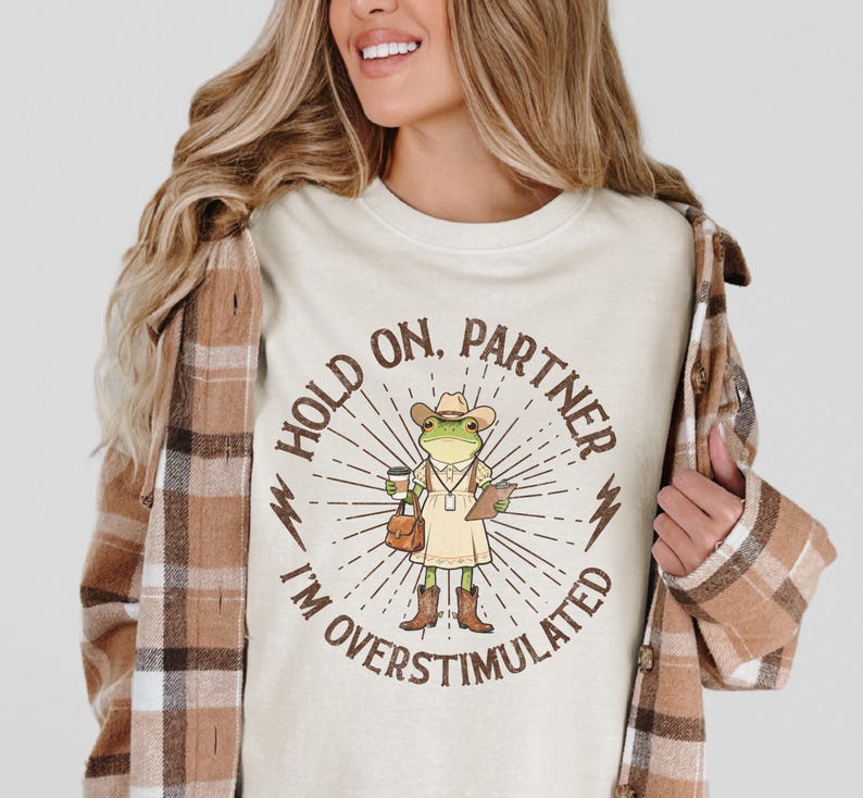 May include: Cream-colored t-shirt featuring a cartoon frog wearing a cowboy hat, holding a coffee cup and clipboard. The text reads "HOLD ON, PARTNER I'M OVERSTIMULATED" in a circular design with sunburst rays and lightning bolts. The shirt is a casual wear item.