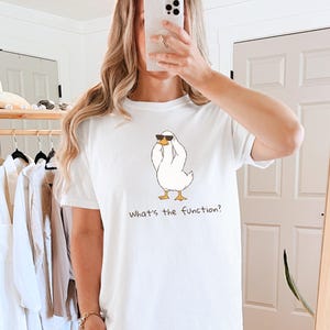 K&ouml;nnte beinhalten: Wei&szlig;es T-Shirt mit einer Cartoon-Ente, die eine Sonnenbrille tr&auml;gt und ihre Augen mit ihren Fl&uuml;geln bedeckt. Der Text "What's the function?" ist darunter gedruckt. Das Shirt ist ein l&auml;ssiger, bequemer Stil.