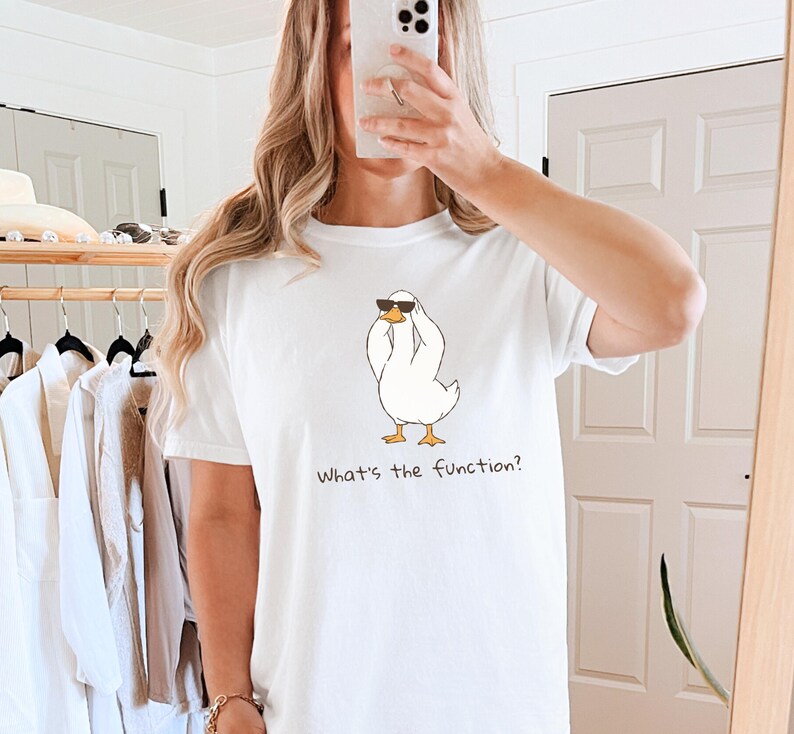 K&ouml;nnte beinhalten: Wei&szlig;es T-Shirt mit einer Cartoon-Ente, die eine Sonnenbrille tr&auml;gt und ihre Augen mit ihren Fl&uuml;geln bedeckt. Der Text "What's the function?" ist darunter gedruckt. Das Shirt ist ein l&auml;ssiger, bequemer Stil.