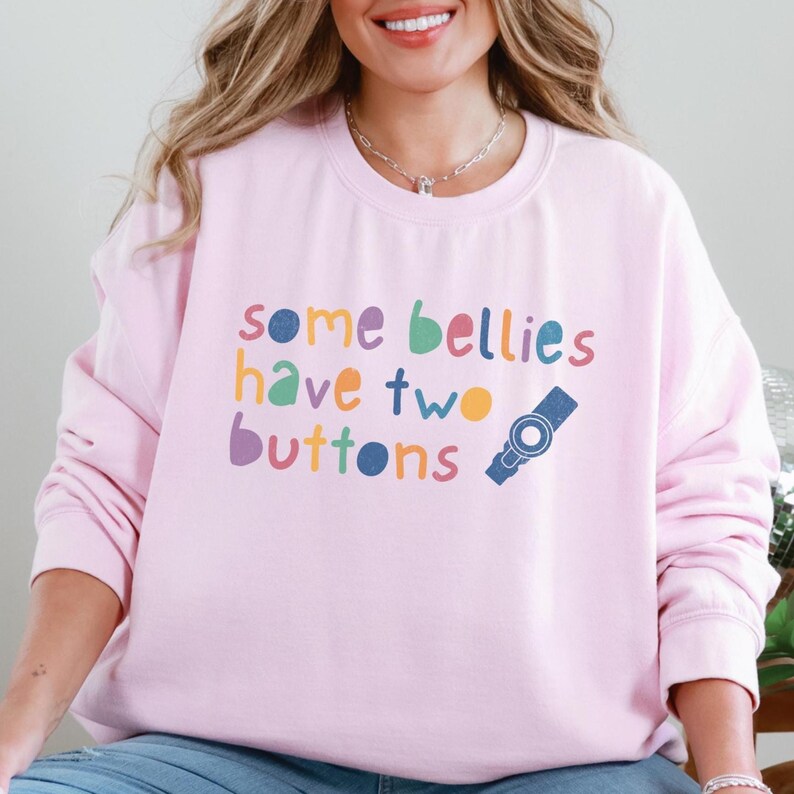 Puede incluir: Sudadera rosa con el texto "some bellies have two buttons" en letras de colores con un gr&aacute;fico de bot&oacute;n azul.