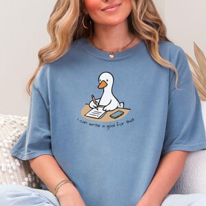 May include: A blue-grey t-shirt featuring a cartoon goose writing on a notepad with a pencil. The goose is sitting at a desk with a smartphone. The text "I can write a goal for that" is below the goose.