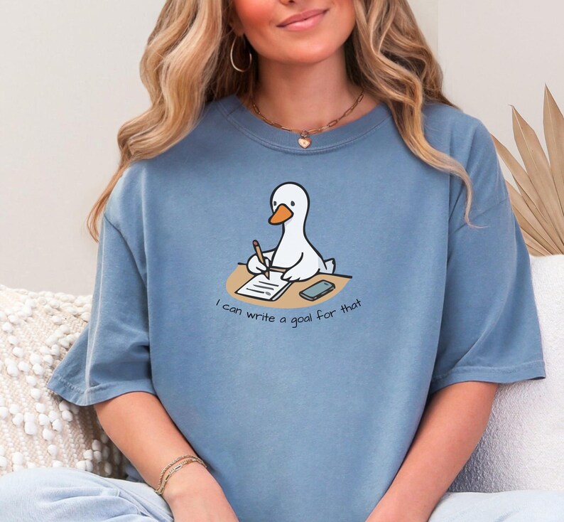 May include: A blue-grey t-shirt featuring a cartoon goose writing on a notepad with a pencil. The goose is sitting at a desk with a smartphone. The text "I can write a goal for that" is below the goose.