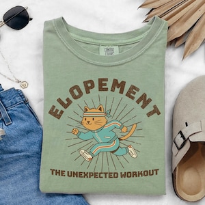 May include: Pale green t-shirt featuring the word "ELOPEMENT" in brown above a cartoon cat in a blue tracksuit and headband. The cat is running with sun rays behind it. The text "THE UNEXPECTED WORKOUT" is below the cat.