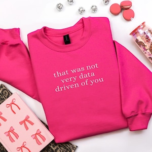 Puede incluir: Una sudadera rosa brillante con el texto blanco "that was not very data driven of you". La imagen incluye elementos decorativos como macarons, un vaso y un cuaderno.