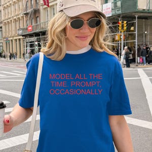 May include: A blue t-shirt with the text "MODEL ALL THE TIME. PROMPT, OCCASIONALLY" in red. The person is wearing a beige cap and sunglasses, holding a white coffee cup with a black lid. The background shows a city street.