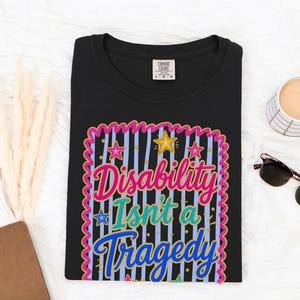 May include: A black t-shirt featuring a graphic design with a pink scalloped border, light blue vertical stripes, and yellow and pink stars. The text reads "Disability Isn't a Tragedy" in pink, teal, and blue script and block lettering.