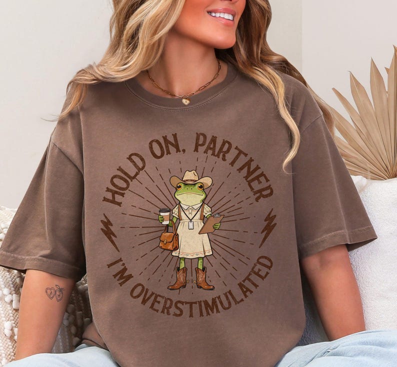May include: Brown t-shirt with a graphic of a frog wearing a cowboy hat, holding a coffee cup and clipboard. Text reads "HOLD ON, PARTNER I'M OVERSTIMULATED" in a circular design. The frog is wearing a dress and cowboy boots.