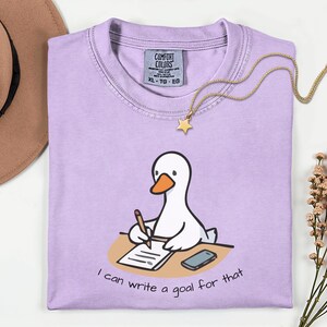 May include: A light purple Comfort Colors XL t-shirt with a cartoon goose writing on a notepad. The text "I can write a goal for that" is below the goose. A gold necklace with a star pendant is on the shirt.