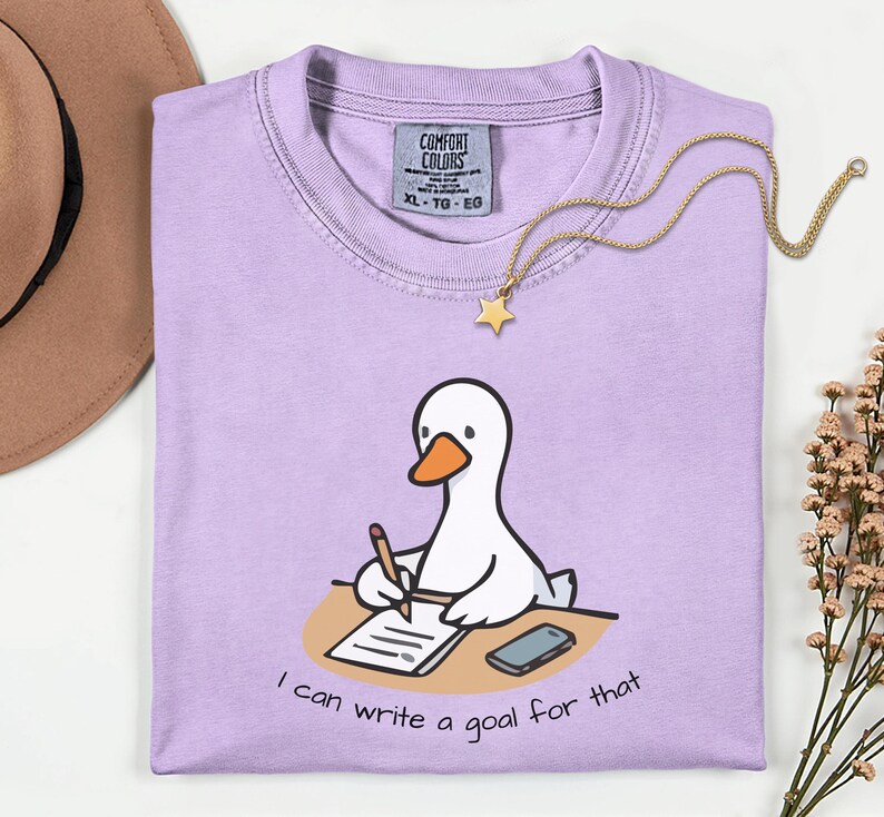 May include: A light purple Comfort Colors XL t-shirt with a cartoon goose writing on a notepad. The text "I can write a goal for that" is below the goose. A gold necklace with a star pendant is on the shirt.