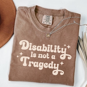 May include: A brown Comfort Colors t-shirt with the text "Disability is not a Tragedy" in a retro font. A silver necklace with a small pendant rests on the shirt. A brown hat is visible in the upper left corner.