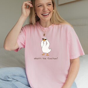 K&ouml;nnte beinhalten: Hellrosa T-Shirt mit einer Cartoon-Ente, die eine Sonnenbrille tr&auml;gt. Die Ente ist wei&szlig; mit gelben F&uuml;&szlig;en und einem schwarzen Schnabel. Der Text "What's the function?" ist darunter gedruckt.