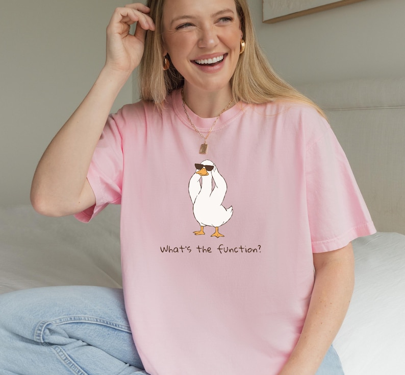 K&ouml;nnte beinhalten: Hellrosa T-Shirt mit einer Cartoon-Ente, die eine Sonnenbrille tr&auml;gt. Die Ente ist wei&szlig; mit gelben F&uuml;&szlig;en und einem schwarzen Schnabel. Der Text "What's the function?" ist darunter gedruckt.