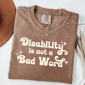 May include: A brown Comfort Colors t-shirt with the text "Disability is not a Bad Word" in a retro font. The shirt has a crew neck and short sleeves. A silver necklace with a pendant is draped across the shirt.