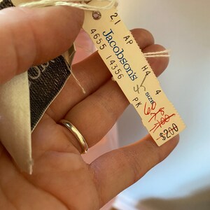 May include: A white tag with black text that reads "Jacobsons" and "4655 14356". The tag is attached to a pink fabric with a string. The tag also has the text "21 AP H4 4 SIZE PK" and a red handwritten price tag that reads "$200".