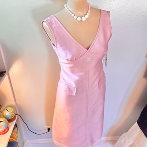 May include: A sleeveless pink dress with a v-neckline and a white pearl necklace. The dress has a small pocket on each side and the letters "RC" embroidered on the front.