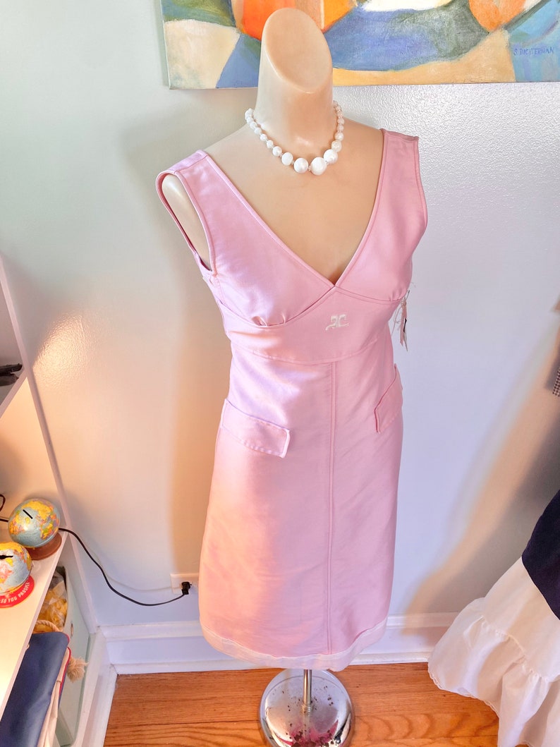 May include: A sleeveless pink dress with a v-neckline and a white pearl necklace. The dress has a small pocket on each side and the letters "RC" embroidered on the front.
