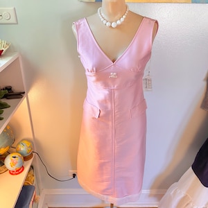 May include: A pink sleeveless dress with a v-neckline and a white pearl necklace. The dress has a small pocket on each side and a white label with the text "Jacqueline" on the right side.
