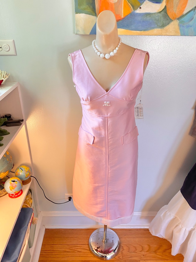 May include: A pink sleeveless dress with a v-neckline and a white pearl necklace. The dress has a small pocket on each side and a white label with the text "Jacqueline" on the right side.