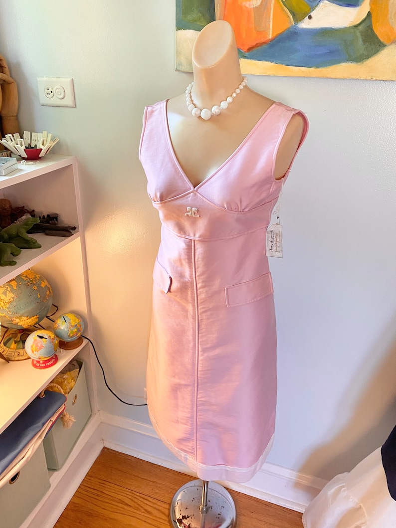 May include: A sleeveless pink dress with a V-neckline and a white pearl necklace. The dress has a small pocket on each side and a white label with the text "Goodwill" on the right side.