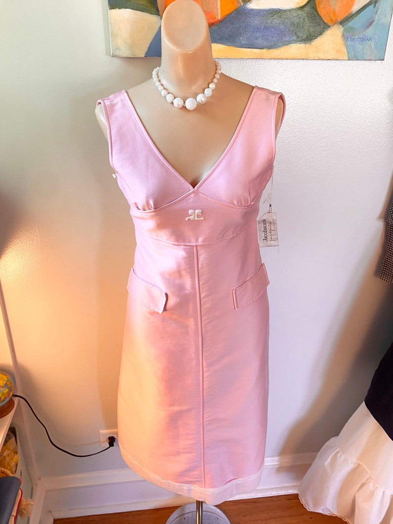 May include: A pink sleeveless dress with a V-neckline and a white pearl necklace. The dress has a small embroidered logo on the front and two pockets on the sides. The dress is on a mannequin.