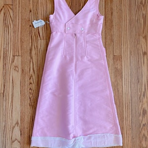 May include: A pink sleeveless dress with a white trim at the hem. The dress has a V-neckline and a button closure at the back. The dress is made of a smooth fabric and has a simple, elegant design.