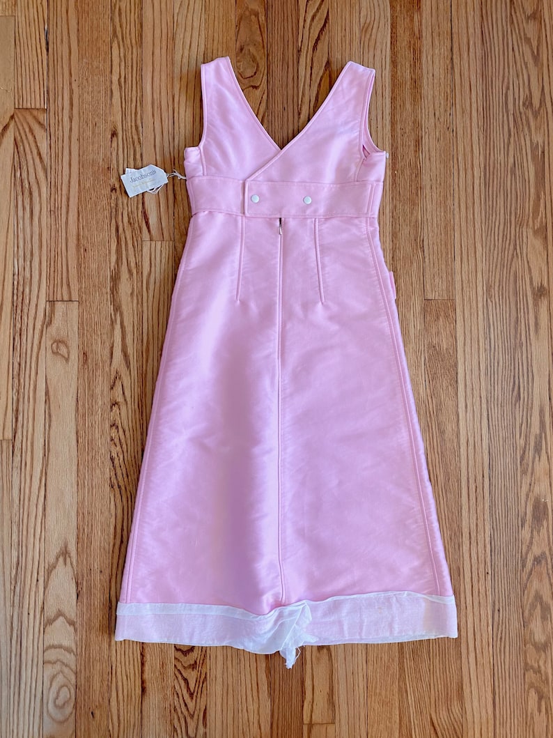 May include: A pink sleeveless dress with a white trim at the hem. The dress has a V-neckline and a button closure at the back. The dress is made of a smooth fabric and has a simple, elegant design.