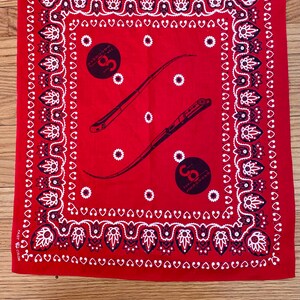 May include: Red bandana with a white and black paisley border. The center features two black illustrations of tools and two black circles with text. The bandana is made of a soft fabric.