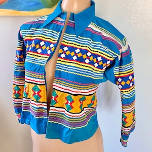 May include: A blue, long-sleeved shirt with a colorful geometric pattern embroidered on the front. The pattern features red, yellow, green, and white accents.