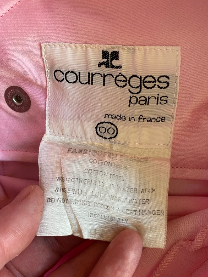 May include: White care label with black text for a pink Courr&egrave;ges Paris garment. The label reads "Fabriqu&eacute; en France", "Cotton 100%", "Wash carefully in water at 40&deg;", "Rinse with luke warm water", "Do not wring, dry on a coat hanger", "Iron lightly".