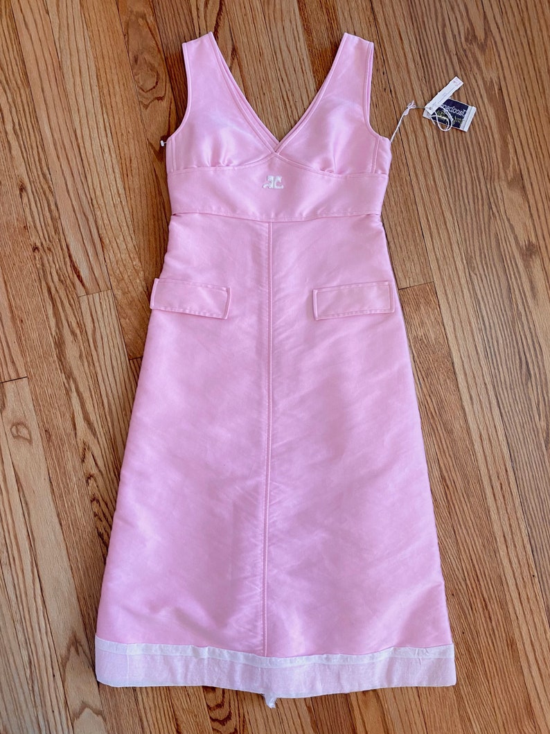 May include: A sleeveless pink dress with a white embroidered logo on the front. The dress has a v-neckline and a white trim at the hem.