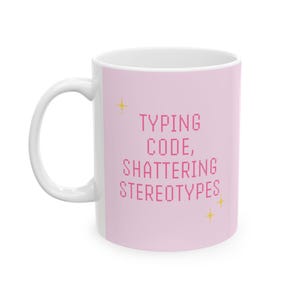 以下が含まれることがあります： ピンクの背景に「TYPING CODE, SHATTERING STEREOTYPES」というピンクの文字が書かれた白いセラミックマグカップ。マグカップには小さな金色の星が描かれている。
