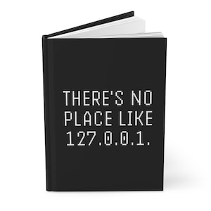 May include: Black journal with the text "THERE'S NO PLACE LIKE 127.0.0.1." in white. The journal is open, revealing white pages. The cover is solid black.