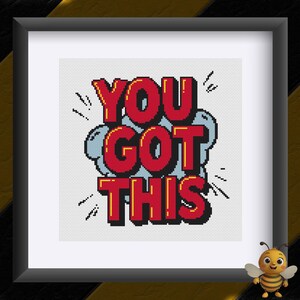May include: Framed cross-stitch artwork with the words "YOU GOT THIS" in red, outlined in yellow, on a white background. The text is in a comic book style, with a blue cloud and black lines for emphasis. The frame is black.