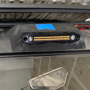 May include: Black LED light bar with amber lights, mounted on a vehicle. A spare tire is mounted above the light bar. The light bar has a black housing and is mounted on the roof of the vehicle.