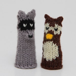 May include: Two knitted finger puppets, one is a raccoon with a black mask and the other is an owl with a yellow beak and white belly.