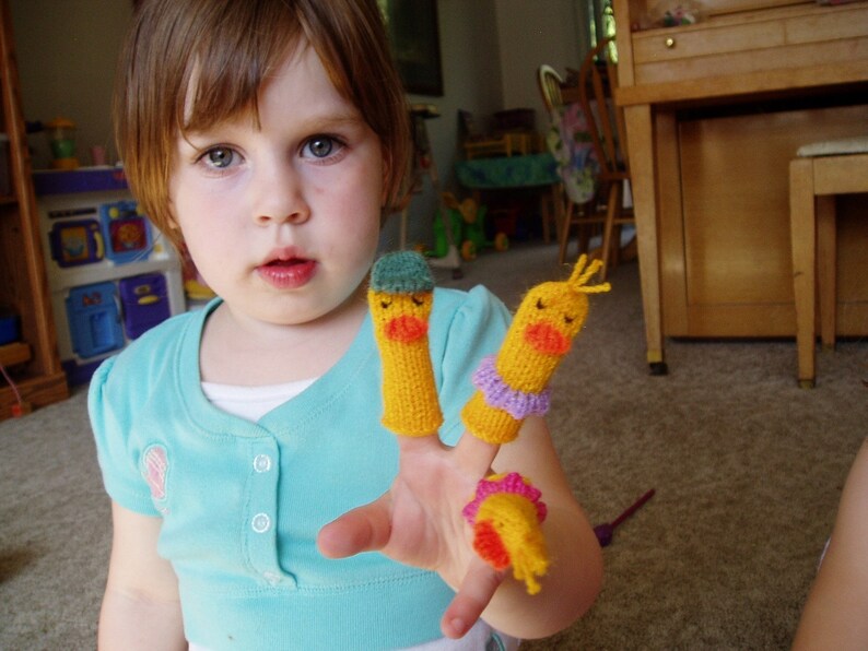 May include: A young person wearing a light blue shirt is playing with three finger puppets. The puppets are yellow with orange beaks and have different colored accents.