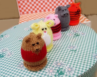 May include: Five knitted animal figurines, each with a different color and design. The figurines are arranged in a row on a green and white checkered tablecloth with floral designs.