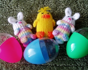 May include: Three knitted Easter toys, a yellow chick, and two white bunnies, with rainbow colored sweaters, are arranged with three plastic Easter eggs, one pink, one blue, and one green.