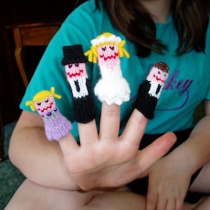 May include: A hand wearing four finger puppets. The puppets are a bride in a white dress, a groom in a black suit, a girl in a purple dress, and a boy in a black suit.